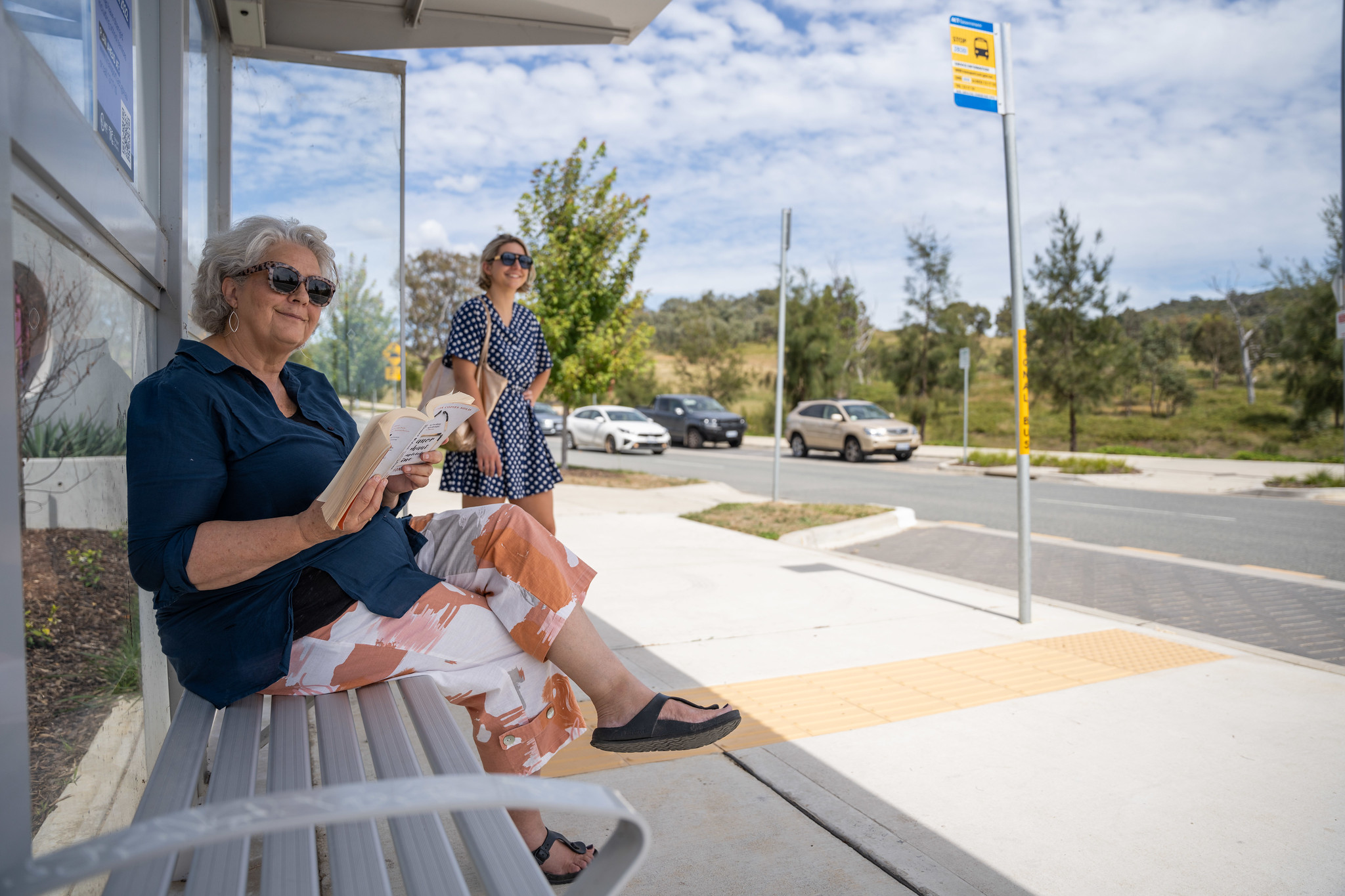 A seated and a standing woman waiting at a covered bus stop. Both are smiling.