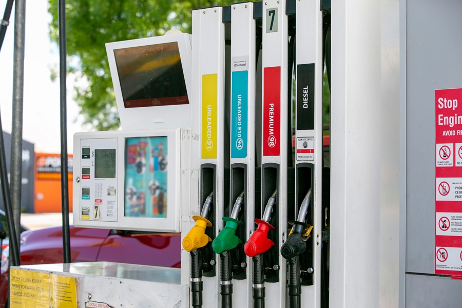 A fuel pump station with four fuel nozzles in different colours—yellow for E10, green for unleaded 91, red for premium 95, and black for diesel—mounted on a white dispenser. A small digital screen and a larger blank display are visible above the pump, with a red car parked in the background.
