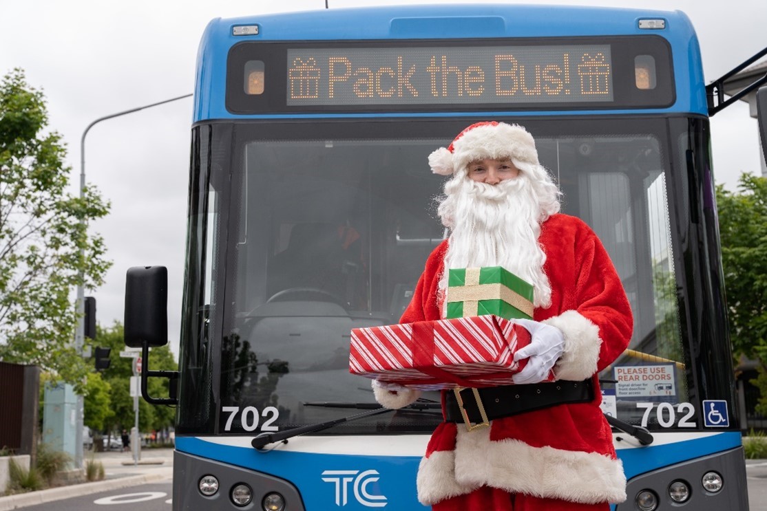 Santa standing in front of a blue bus hold a present.